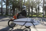 Retro motorbike with installed surfboard rack carrying white surfboard on palm-lined beach street, real-world surf carrier usage demo